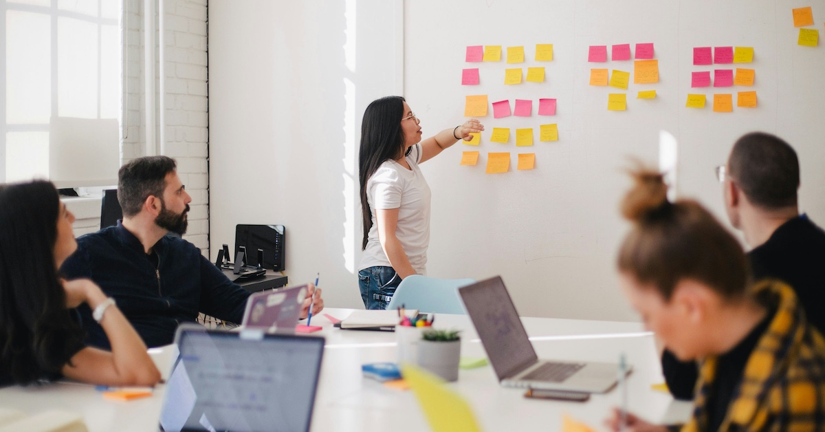 Mennesker i et møterom, som følger med på en kvinne som henger post-it lapper på en vegg.